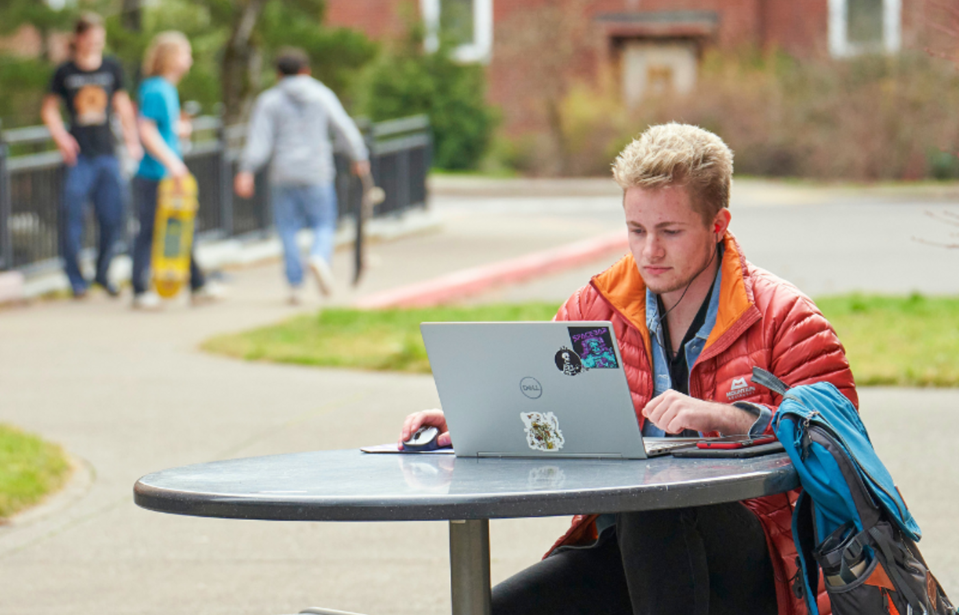Male college student using laptop at an outdoor table on campus with other students in the background