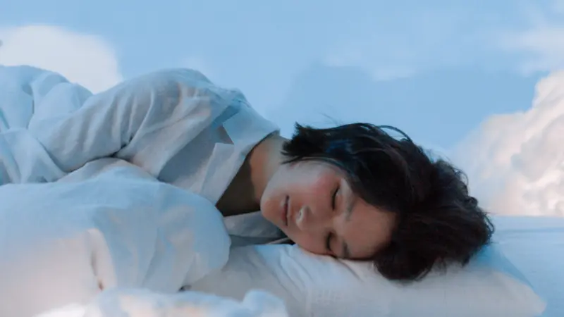 A young adult woman sleeping with her head on a pillow in a calm blue space with fluffy faux clouds surrounding her