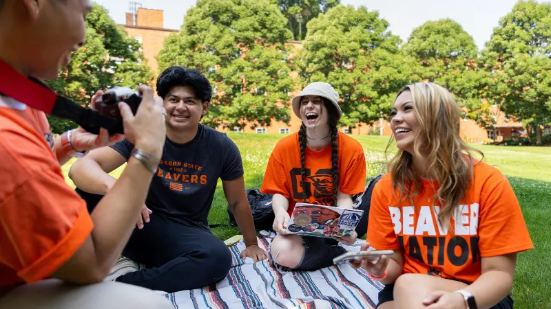 Four students sitting together on a blanket in the MU Quad, chatting and laughing