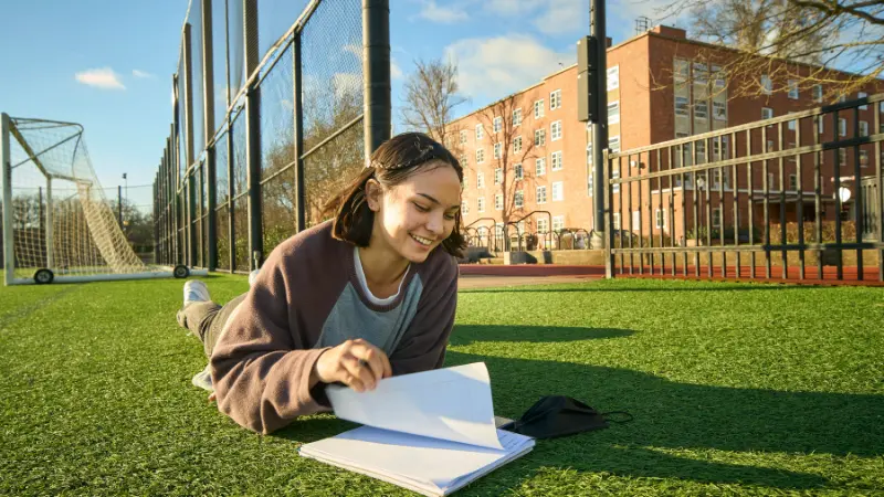 A female-appearing student laying on her stomach in the grass of sports field doing school work