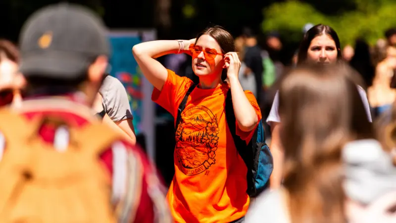Student wearing an OSU t-shirt and orange heart shaped sunglasses standing outside on campus surrounded by other students walking by