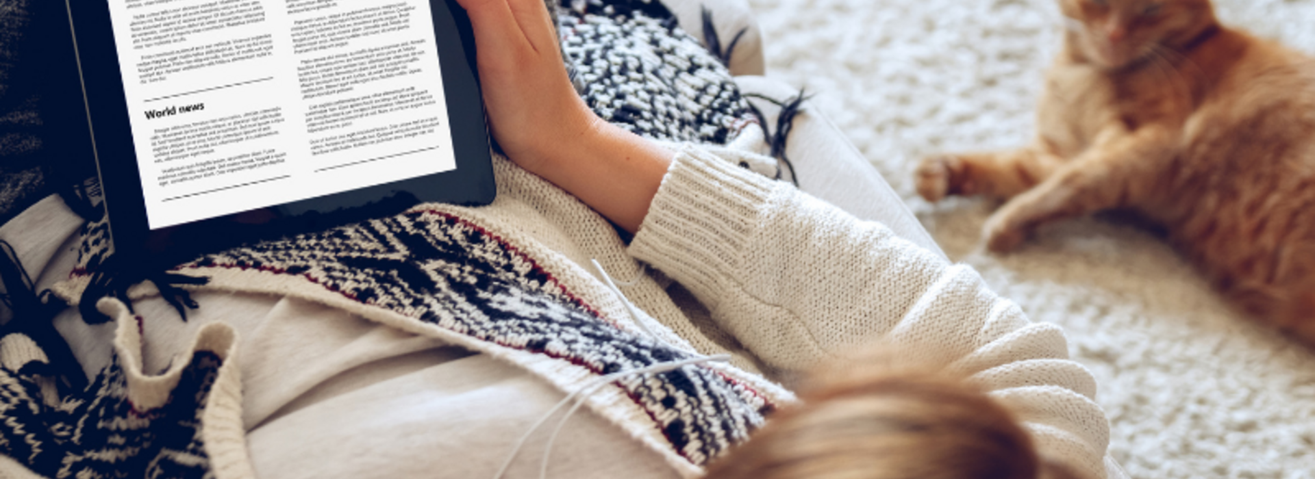 A young woman sitting on the floor reading the news on a tablet with a cat laying next to her