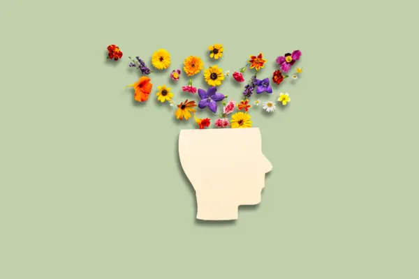 A beige silhouette of a human head is open at the top, with colorful flowers of various shapes and sizes emerging from it, set against a soft green background. 