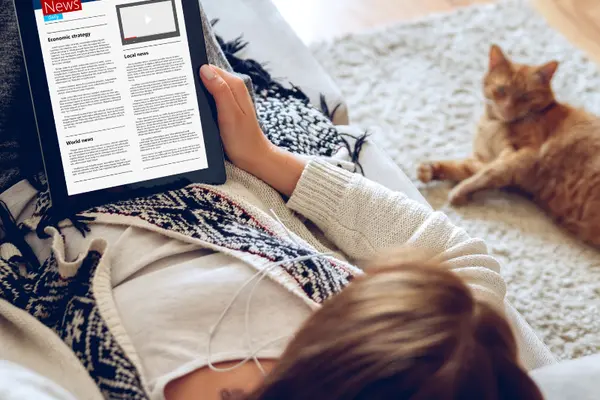 A young woman sitting on the floor reading the news on a tablet with a cat laying next to her